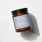 Amber jar candle with a blue label that reads "Kiss the Brides VOWED" in pink script.