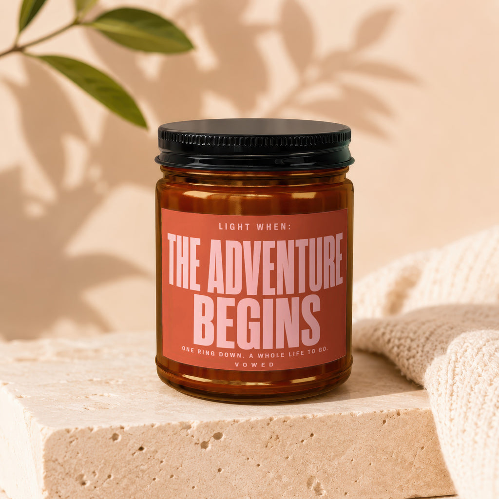Amber candle with black lid in a jar, labeled "THE ADVENTURE BEGINS".