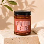 Amber candle with black lid in a jar, labeled "THE ADVENTURE BEGINS".