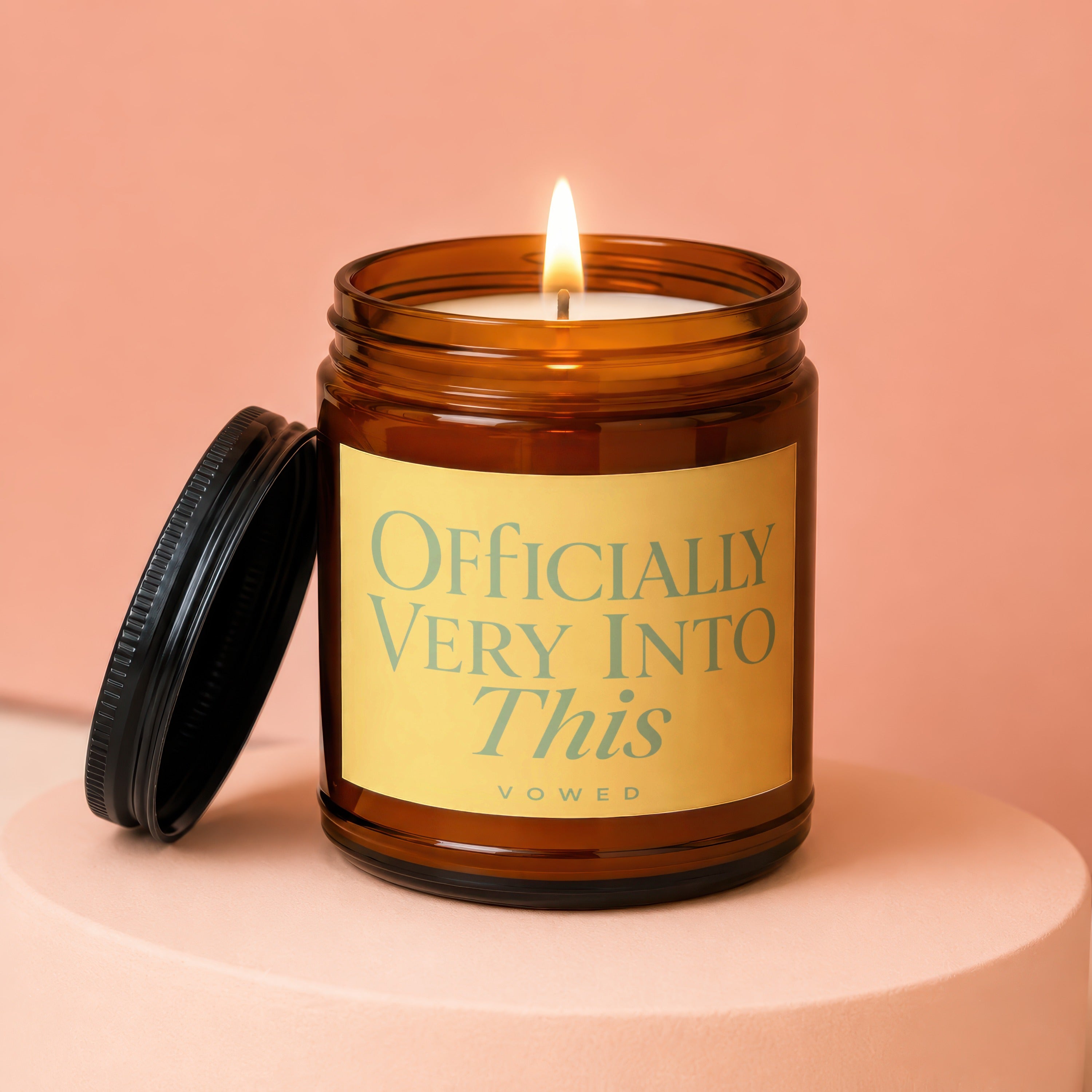 Amber glass candle with lit wick, text "OFFICIALLY VERY INTO This vowed" on label.