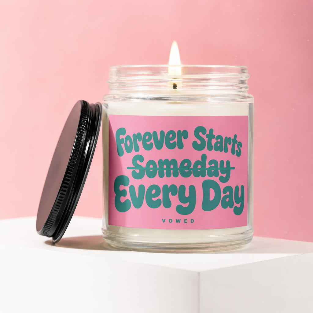 Lit candle in glass jar with pink label reading "Forever Starts Someday Every Day".