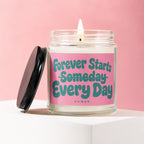 Lit candle in glass jar with pink label reading "Forever Starts Someday Every Day".