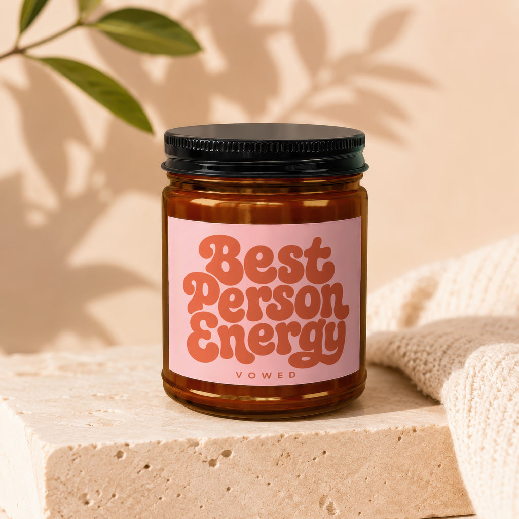 Amber candle with 'Best Person Energy' text on pink label, with shadows and knit fabric.