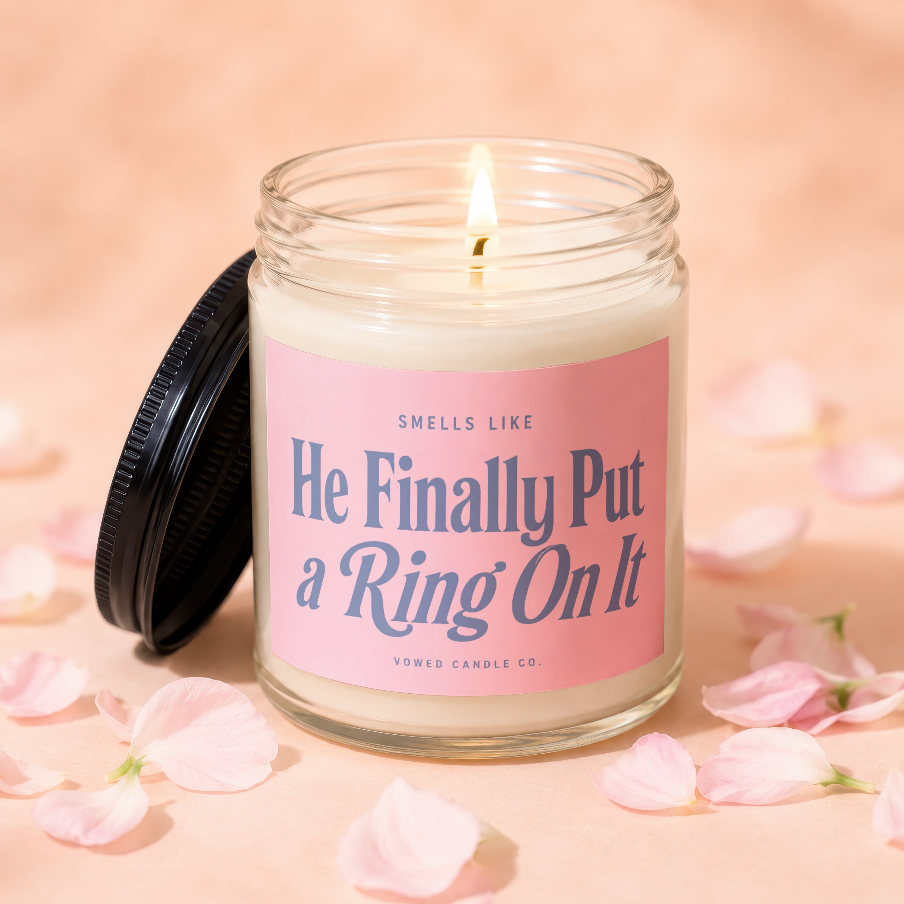 Glass candle with lit wick and pink label that reads "He Finally Put a Ring On It".