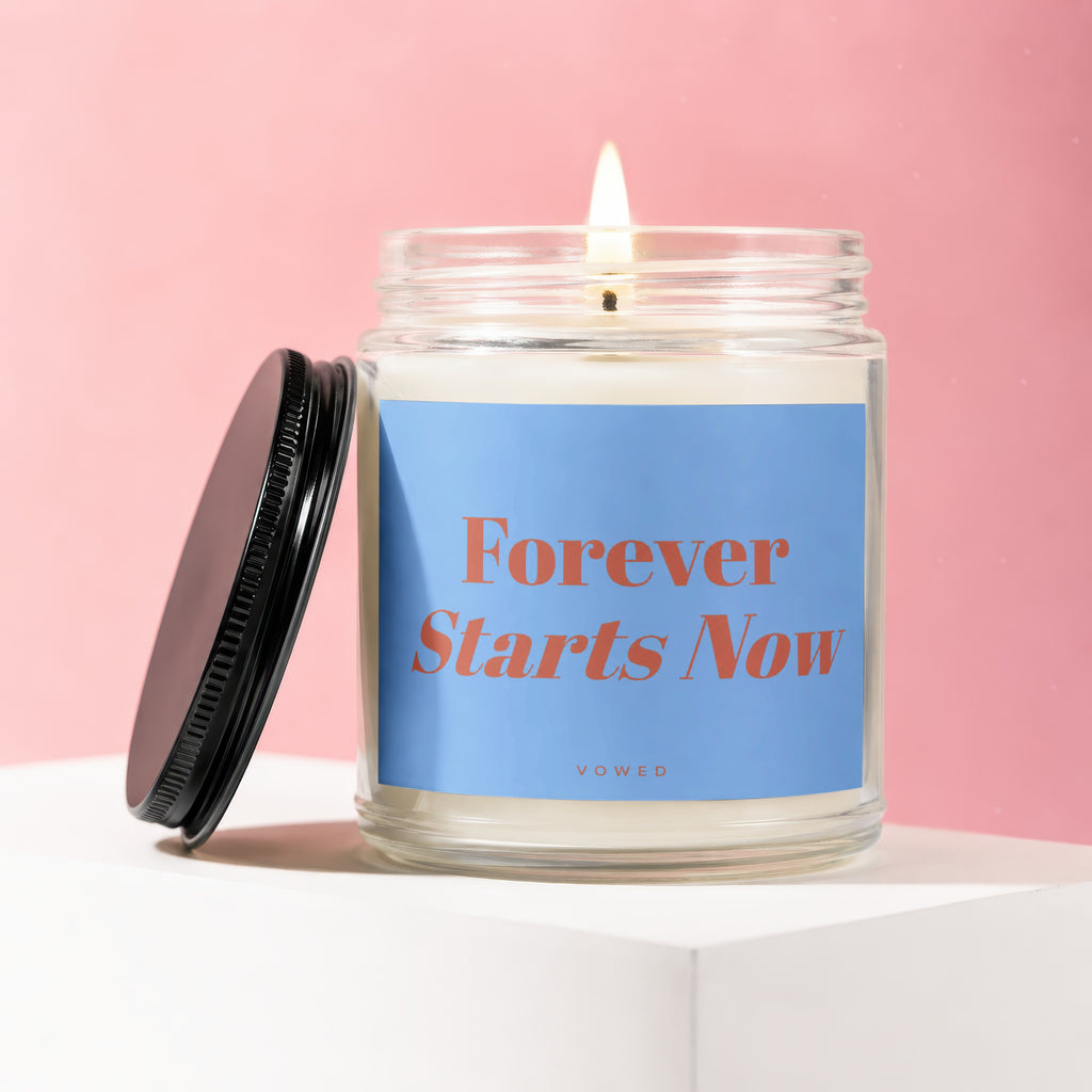 Candle with illuminated wick and blue label reading "Forever Starts Now VOWED".