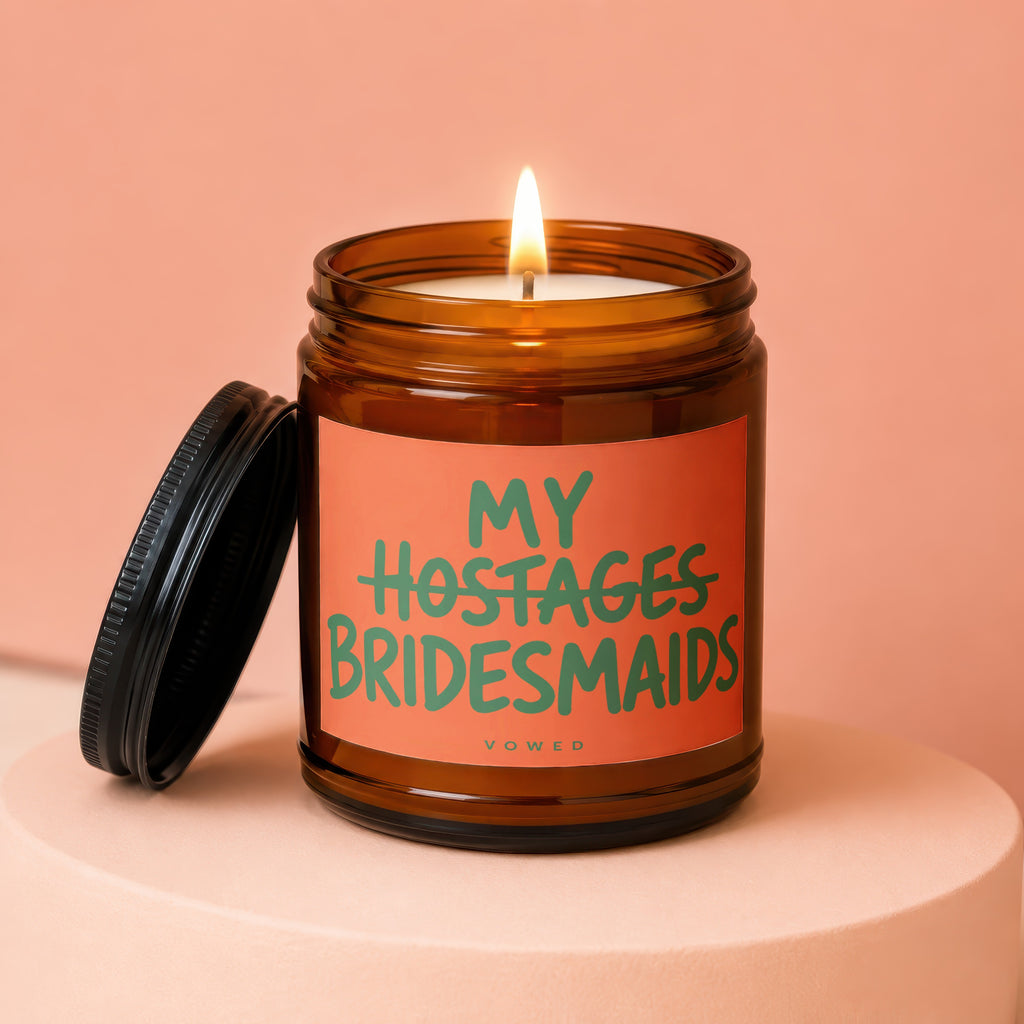 Amber jar candle with lit wick and a label reading "MY HOSTAGES BRIDESMAIDS" VOWED.
