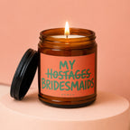 Amber jar candle with lit wick and a label reading "MY HOSTAGES BRIDESMAIDS" VOWED.