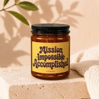 Amber jar candle with yellow label reading "Mission Impossible Accomplished Vowed".