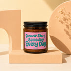 Amber jar candle with black lid, pink label reading "Forever Starts Someday Every Day"