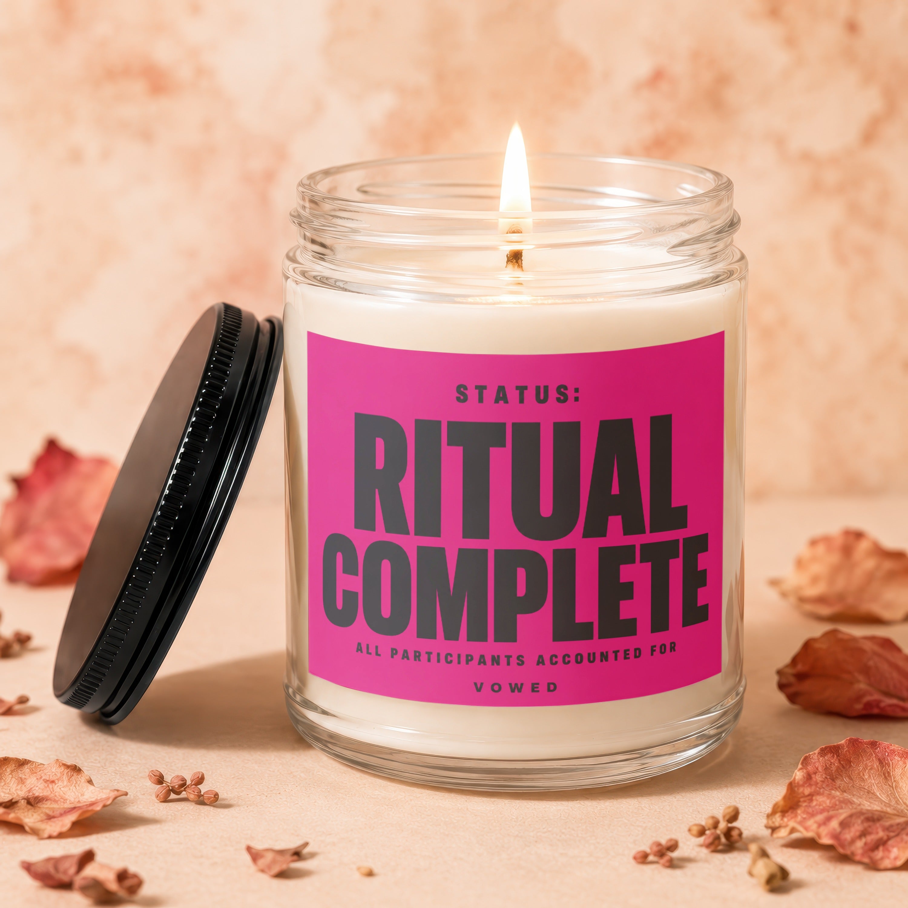 Jar candle with lit wick and pink label reading "STATUS: RITUAL COMPLETE".