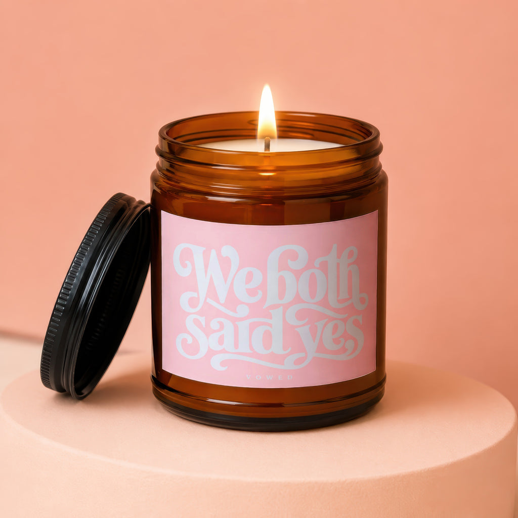 Amber glass candle with lit wick and "We both said yes" on pink label.