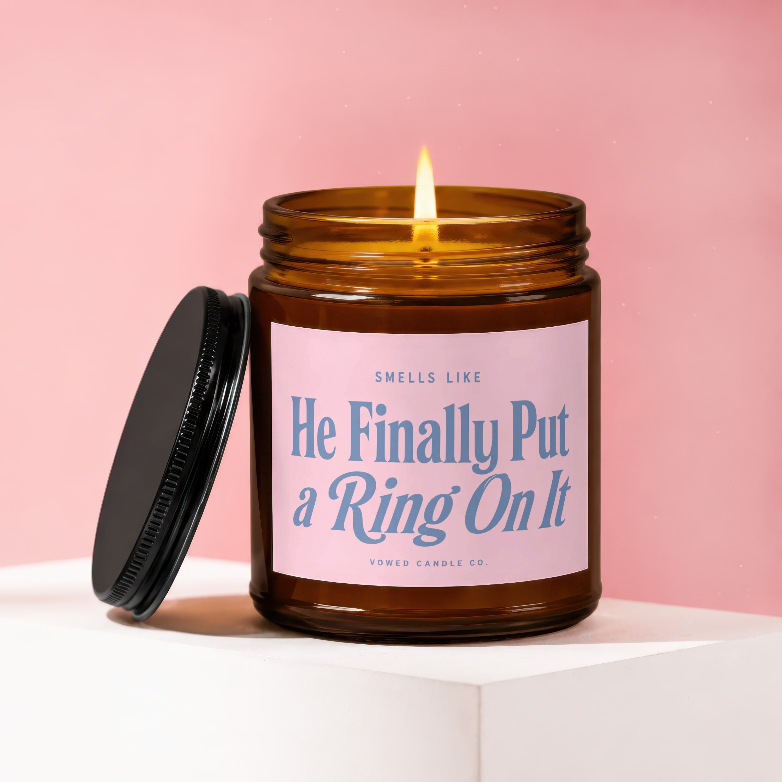 Amber candle with lit wick and pink label reading "SMELLS LIKE He Finally Put a Ring On It"