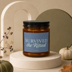 Amber glass candle with blue label reading "Survived the Ritual" and "vowed."