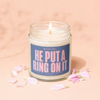 Lidded glass jar candle with text "WARNING: HE PUT A RING ON IT" and "SHE HAS NOT STOPPED LOOKING AT IT".