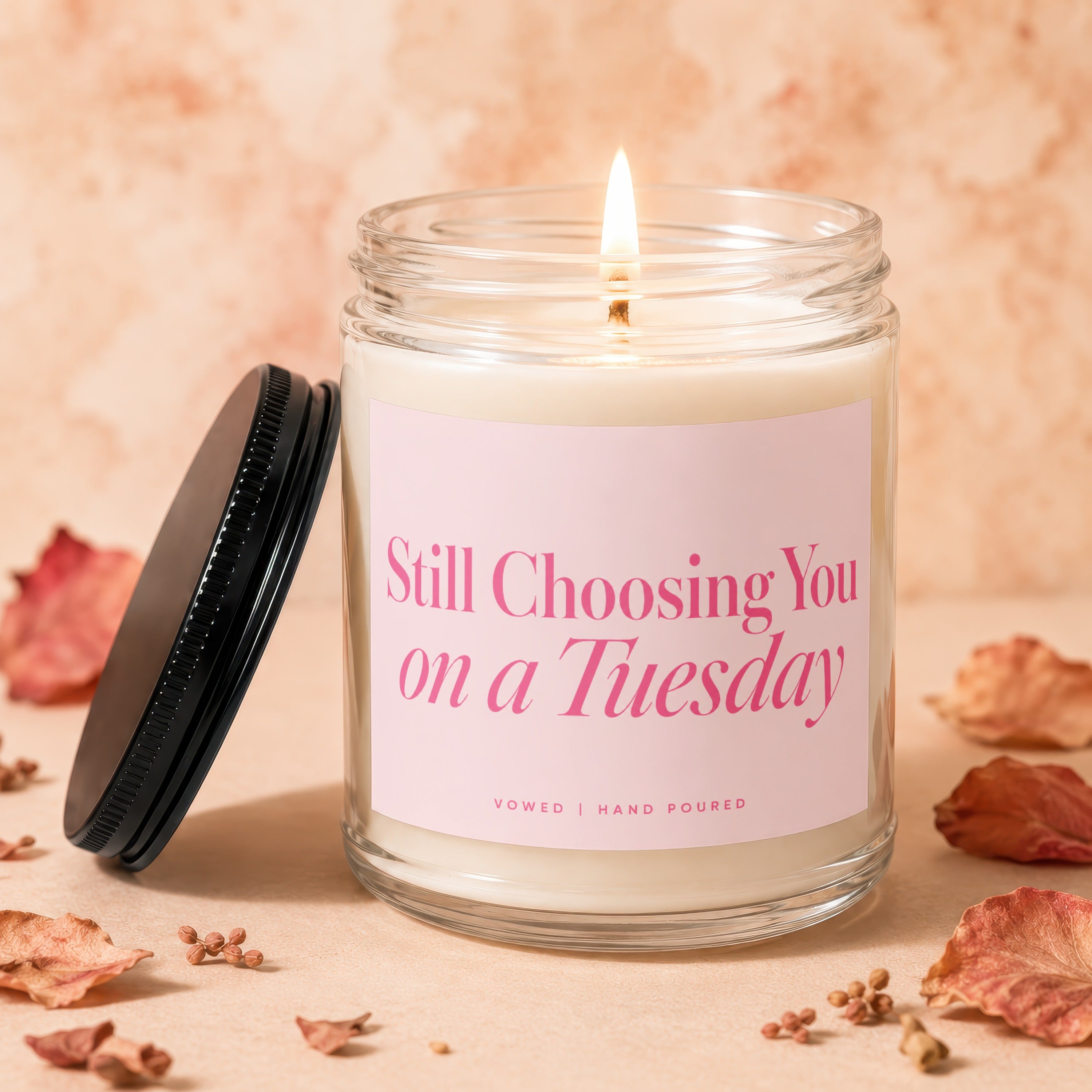 Glass candle with lit wick, pink label reading "Still Choosing You on a Tuesday."