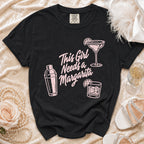 Black Comfort Colors t-shirt with pink "This Girl Needs a Margarita" graphic and drink icons.