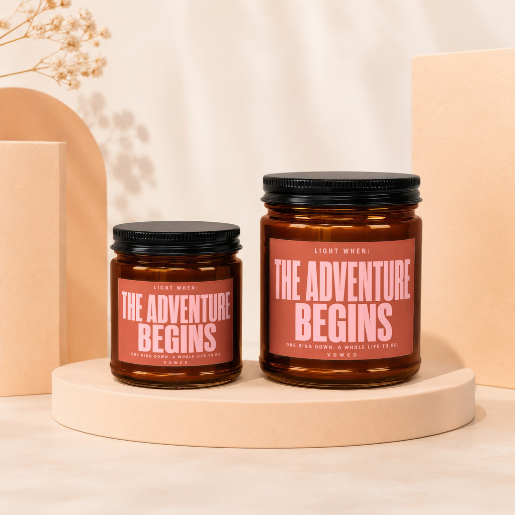 Two amber glass candles with "The Adventure Begins" printed on their labels.