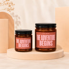Two amber glass candles with "The Adventure Begins" printed on their labels.