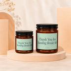 Two amber glass candles with black lids and light blue labels reading "Thank You for Standing Beside Me".