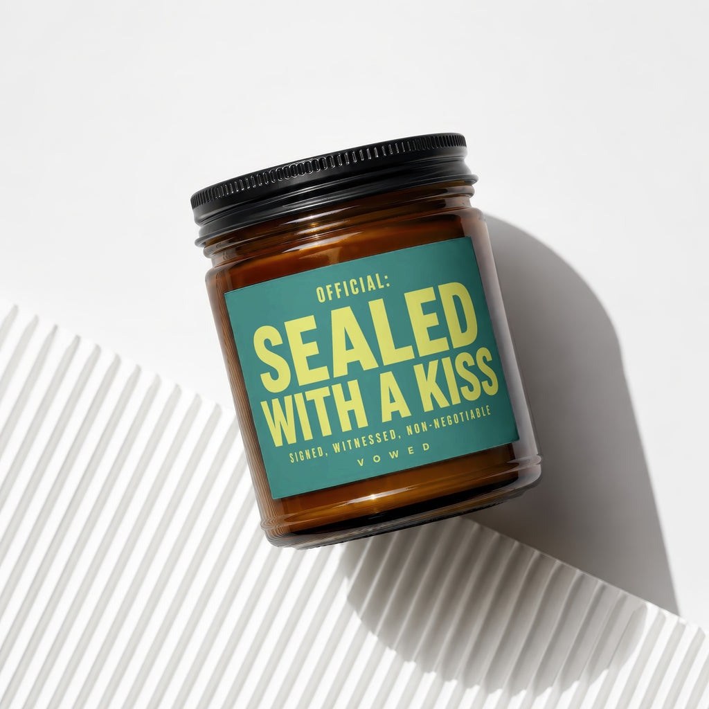 Glass candle with teal label reading "OFFICIAL: SEALED WITH A KISS"