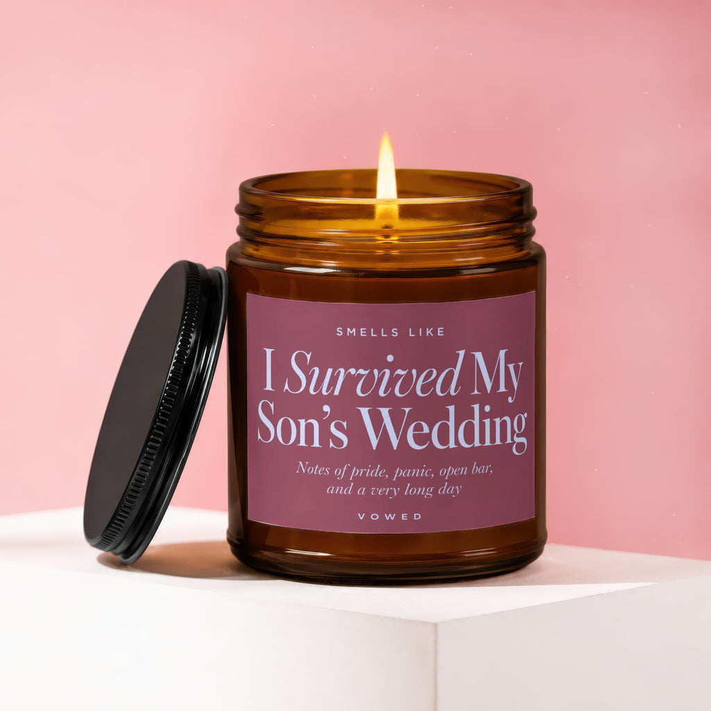 Amber candle with lit wick on pink background, label reads "I Survived My Son's Wedding."