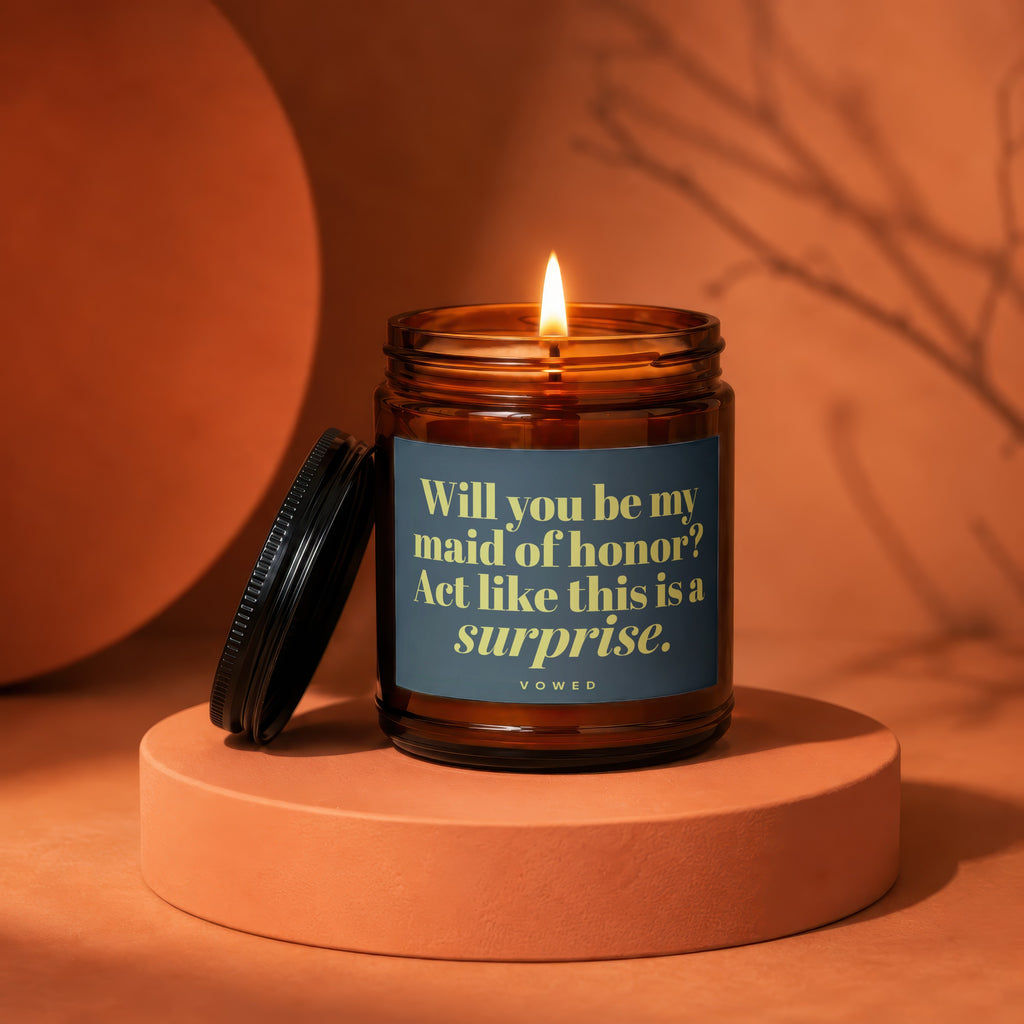 Amber candle with lit flame and dark lid, reading "Will you be my maid of honor? Act like this is a surprise."