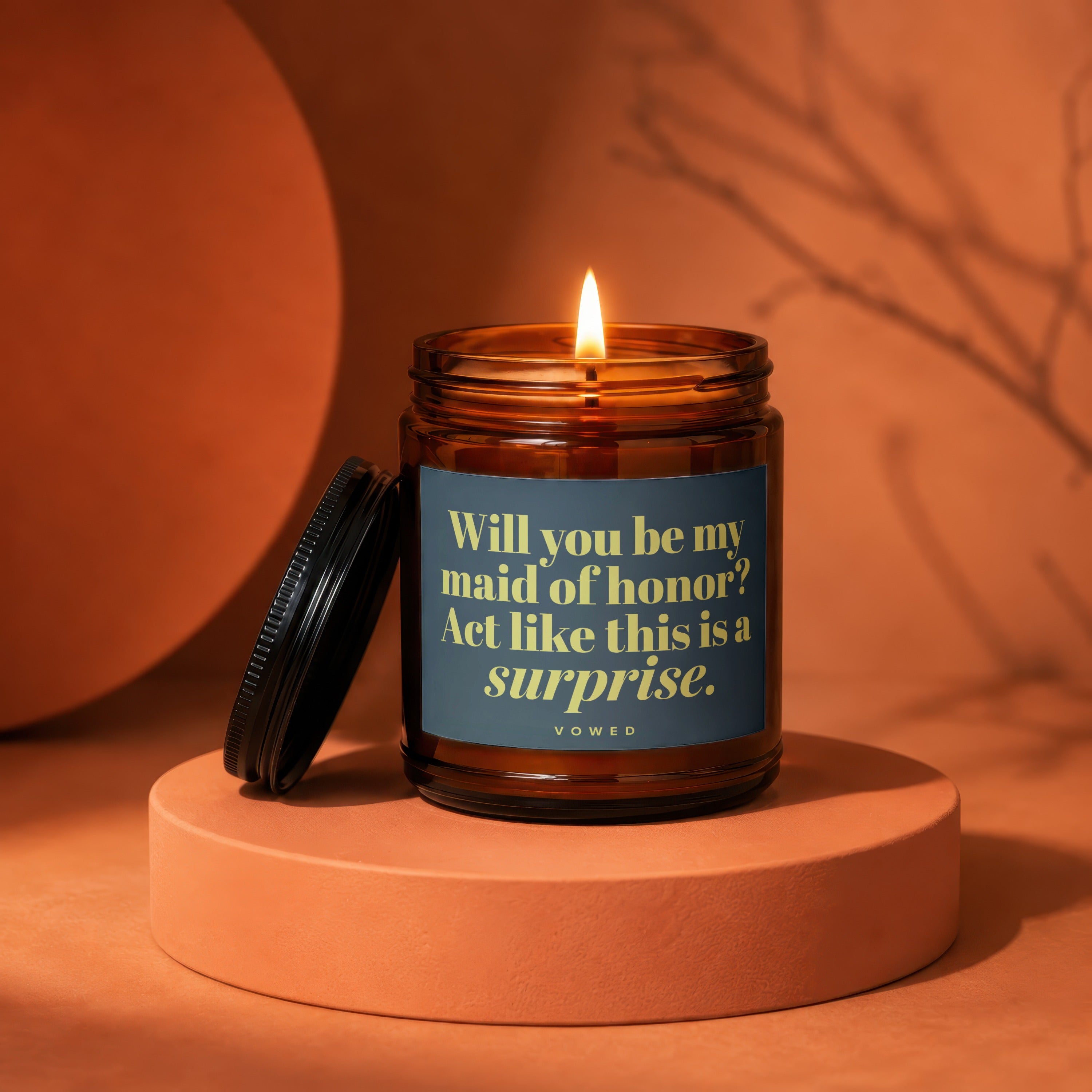 Amber candle with lit flame and dark lid, reading "Will you be my maid of honor? Act like this is a surprise."