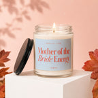 Scented candle with "Mother of the Bride Energy" text, lid off, lit wick.