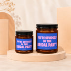 Two amber glass candles with black lids and blue labels reading "You're Obviously In The Bridal Party".