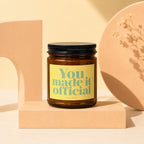 Brown candle with black lid on peach pedestal, label reads "You. made it official."