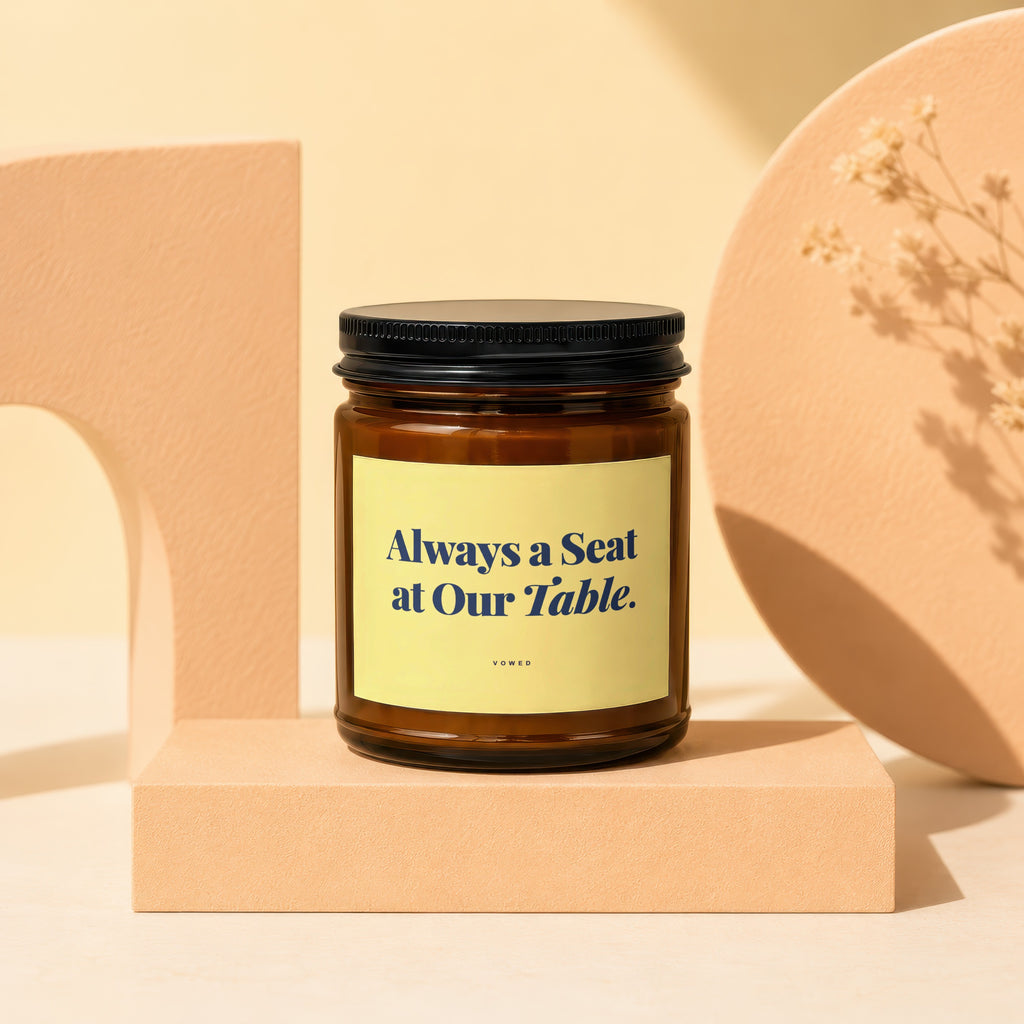 Amber jar candle with black lid, yellow label reading "Always a Seat at Our Table."