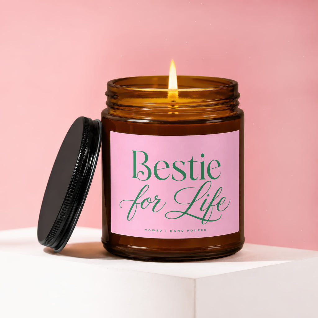 Brown glass candle with lit wick, pink label reads "Bestie for Life," black lid askew.