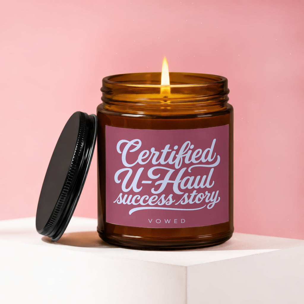 Brown glass candle with lit flame and lid off, displaying text "Certified U-Haul success story".