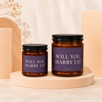 Two amber glass candles with black lids, featuring purple labels asking "Will You Marry Us?"