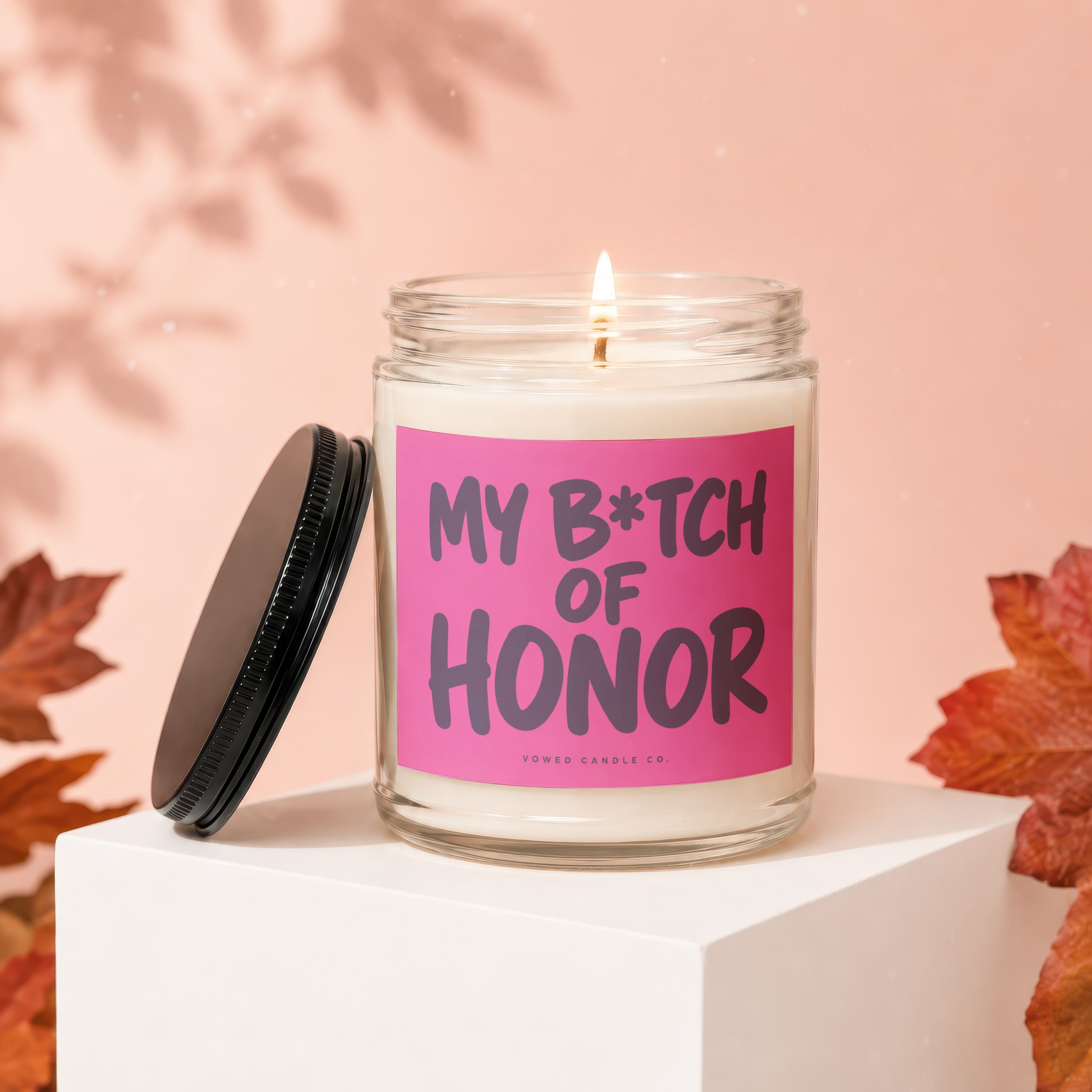 Candle with "My B*tch of Honor" text on a pink label, lid off, autumn leaves.