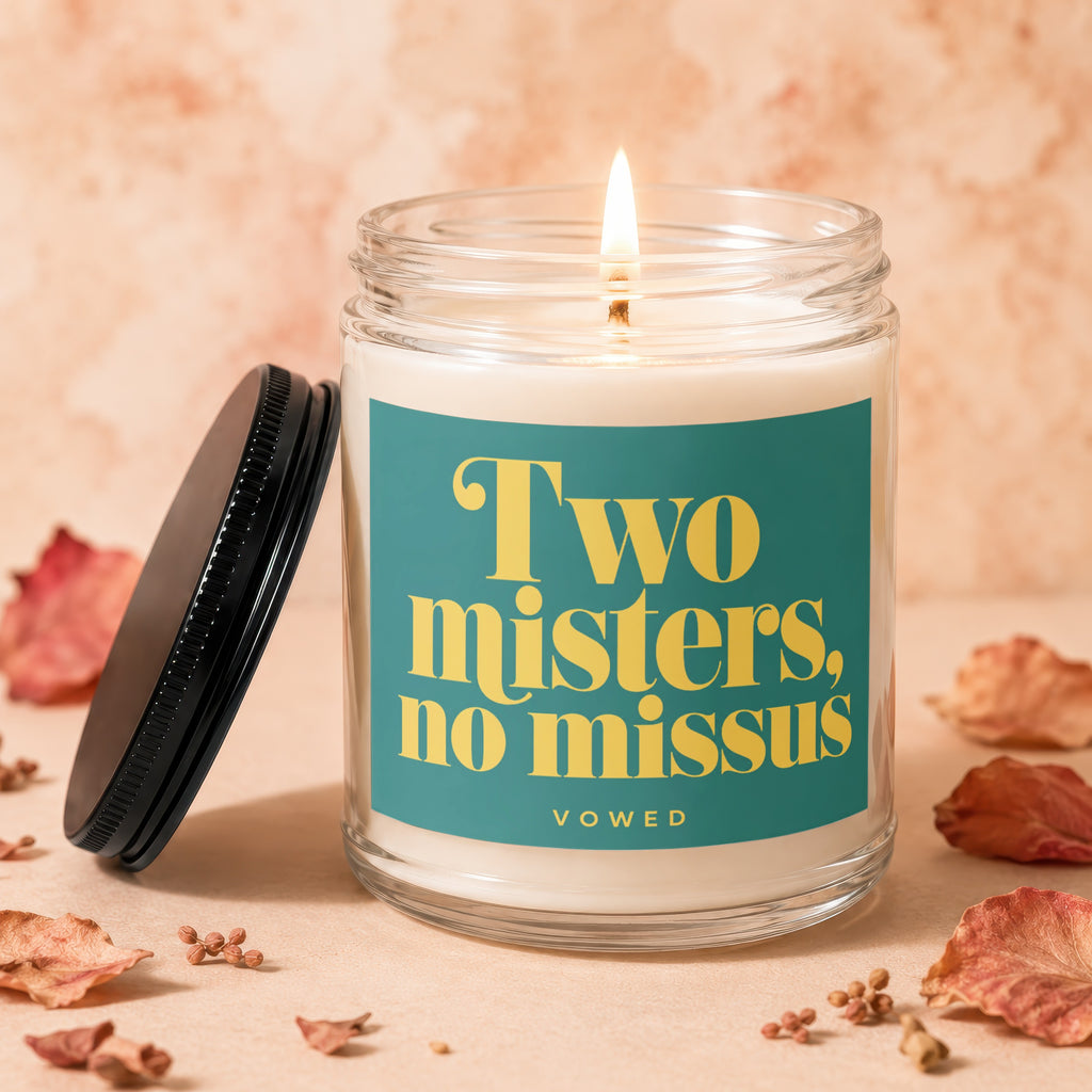 Glass jar candle with lit wick and teal label reading "Two misters, no missus
