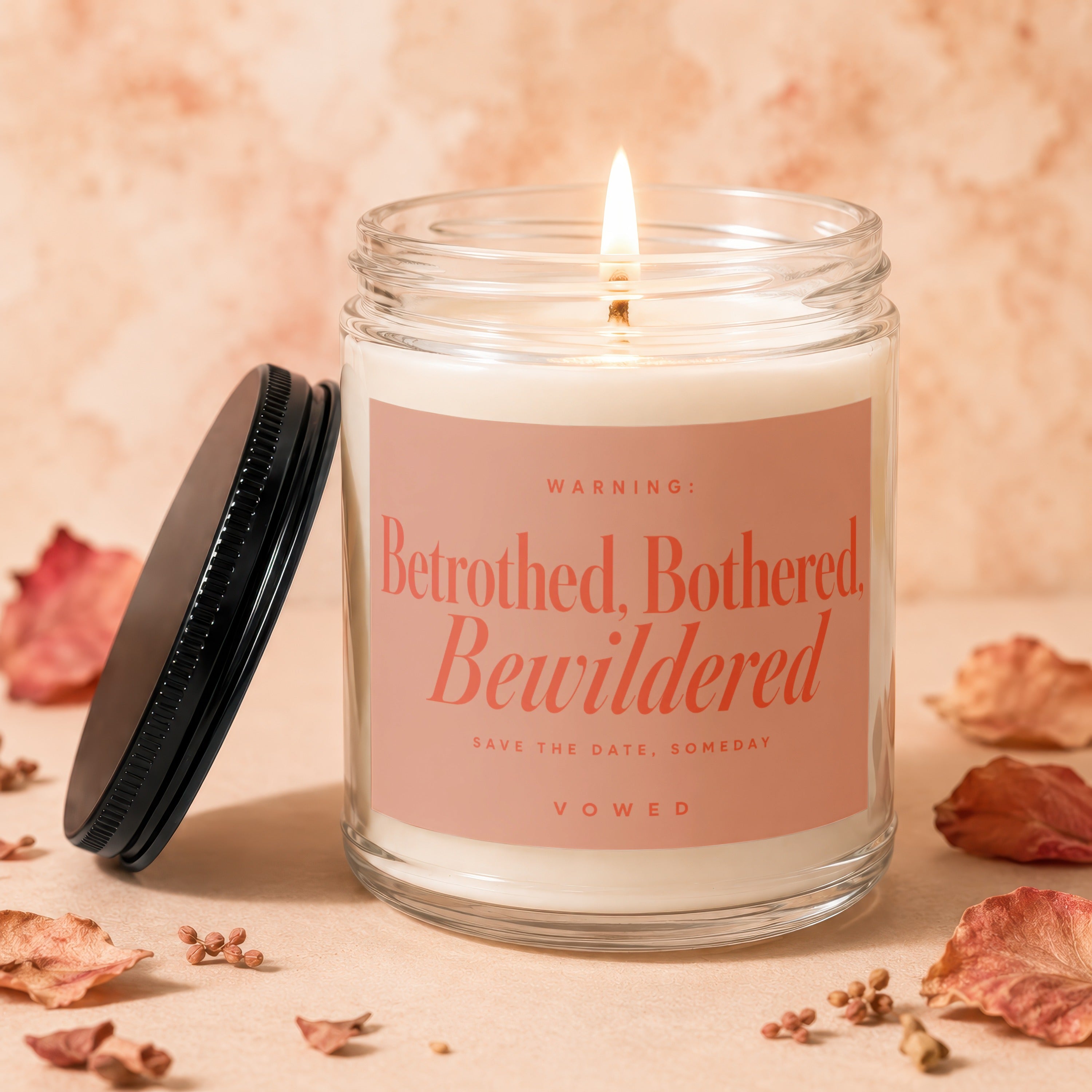 Glass candle with lit wick, featuring "Betrothed, Bothered, Bewildered" text.