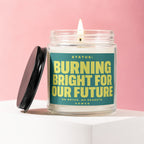 Glass candle with burning wick and teal label reading "Burning Bright for Our Future".