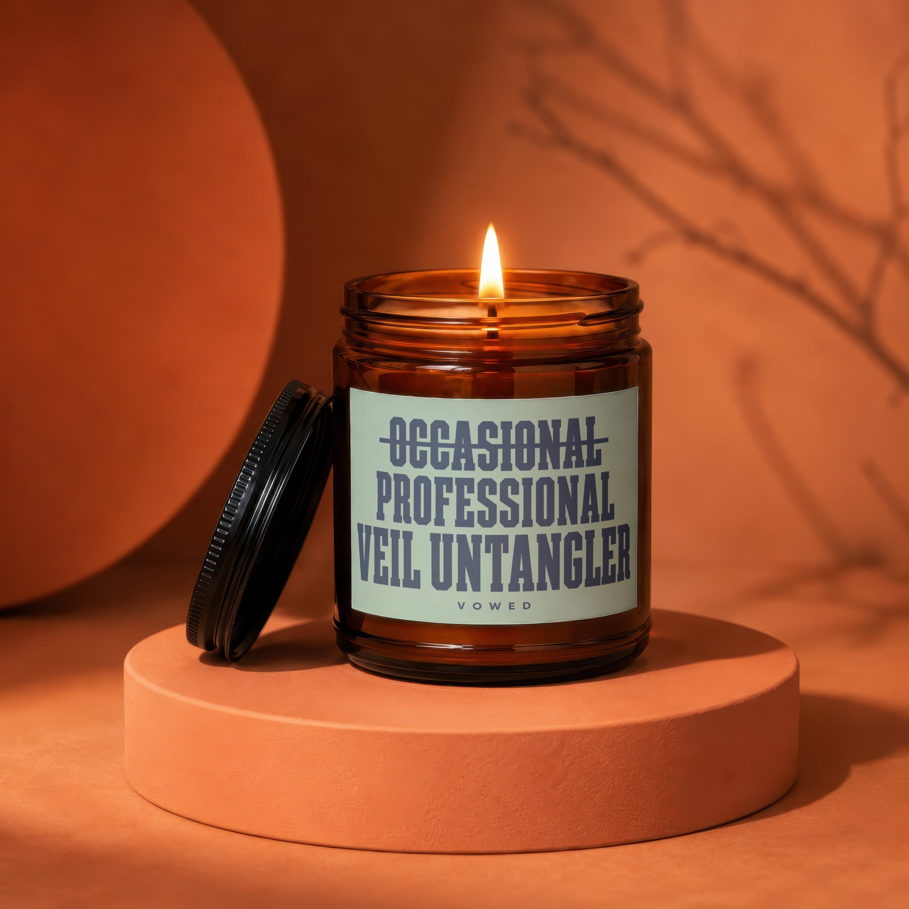 Brown jar candle with burning wick, reads "OCCASIONAL PROFESSIONAL VEIL UNTANGLER".