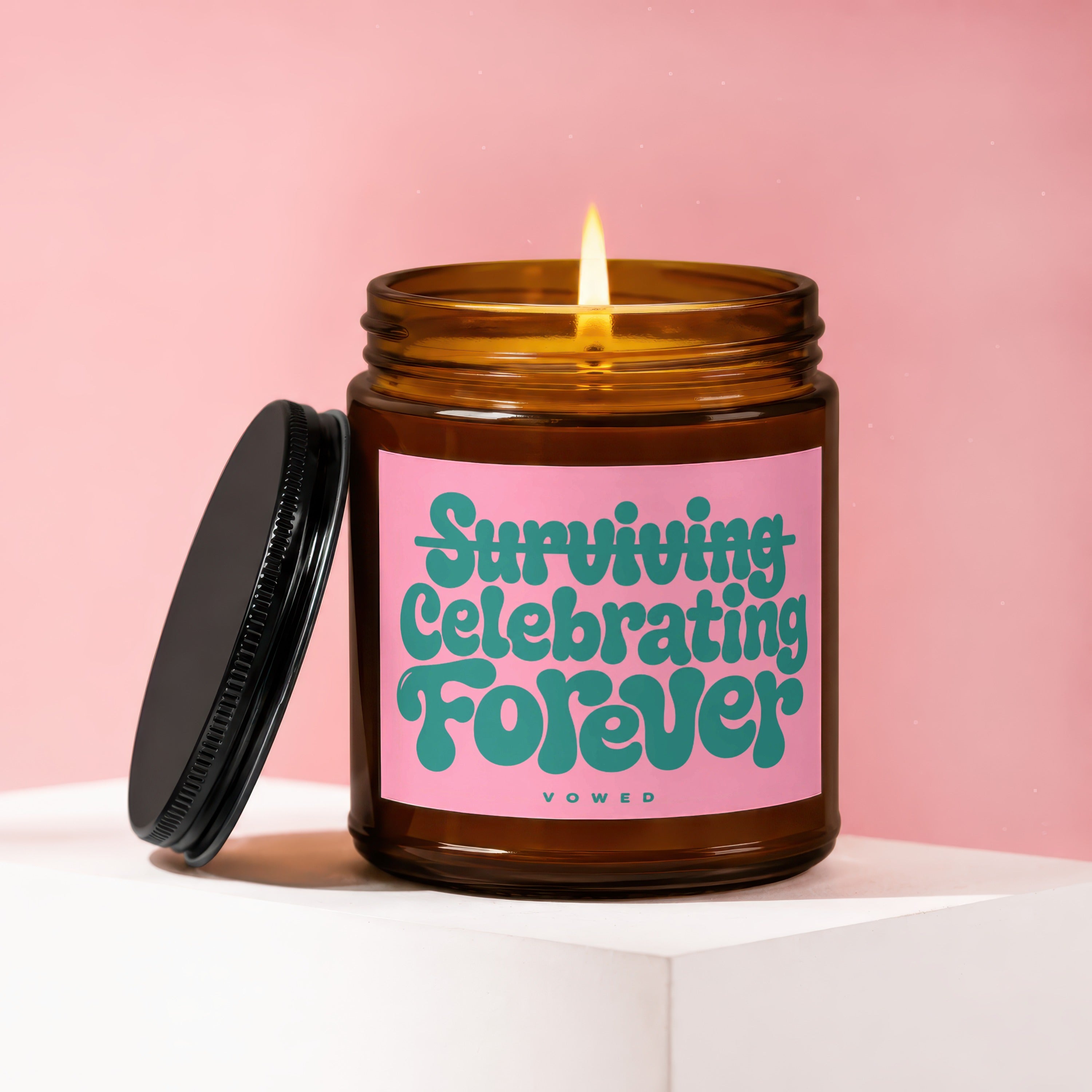 Amber glass candle with lit flame and lid, labeled "Surviving Celebrating Forever"