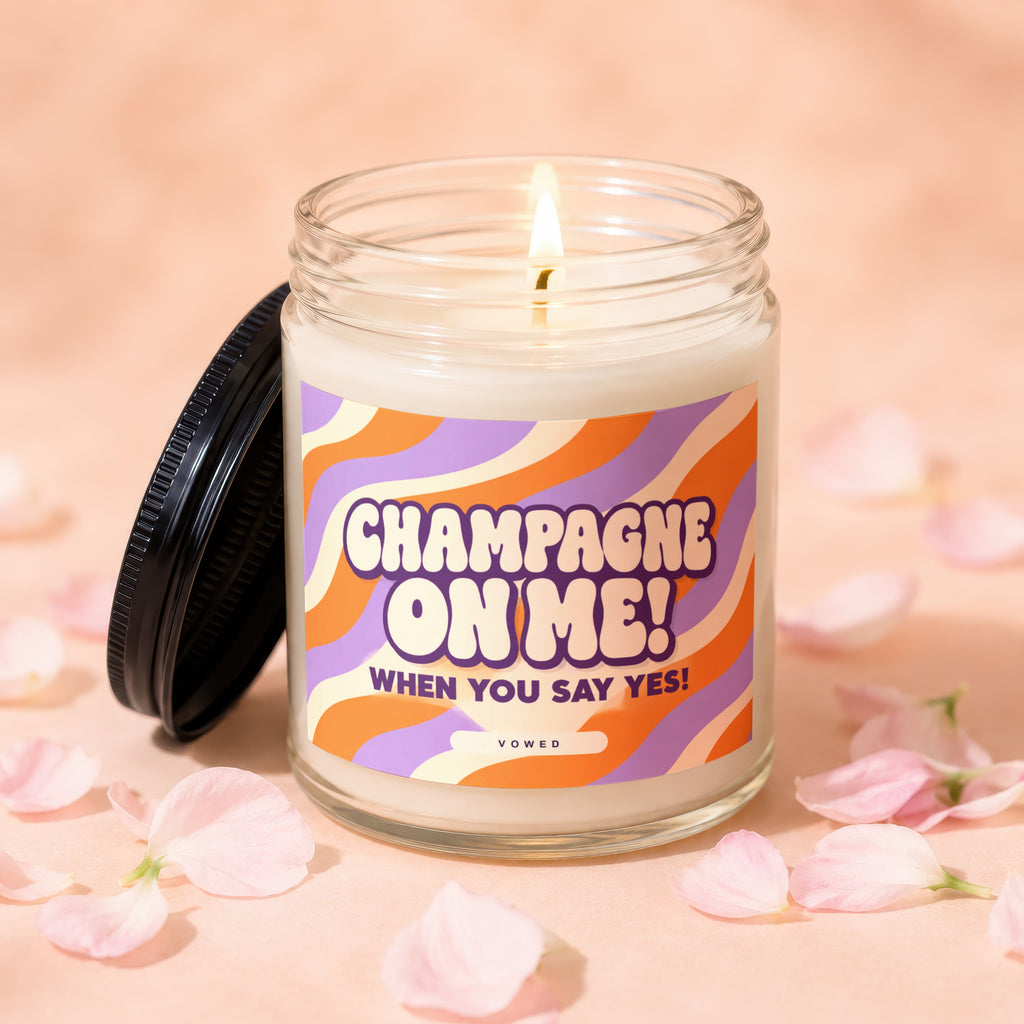 Glass candle with lit wick and a lid, featuring "Champagne on Me! When You Say Yes!" text.