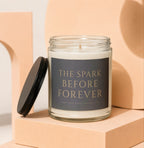 Glass candle with "The Spark Before Forever" label, lid off, on a peach surface.