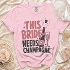 Pink t-shirt with "THIS BRIDE NEEDS CHAMPAGNE" text and champagne bottle graphic.
