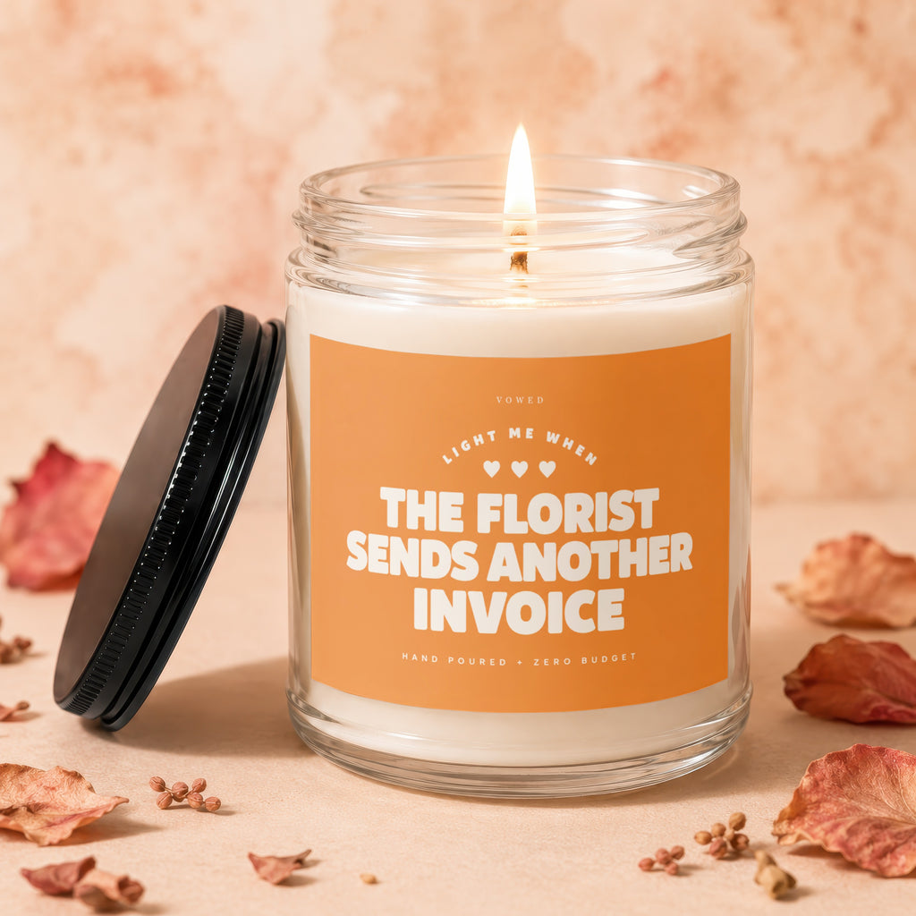 Glass candle with burning wick and orange label reading "The Florist Sends Another Invoice".