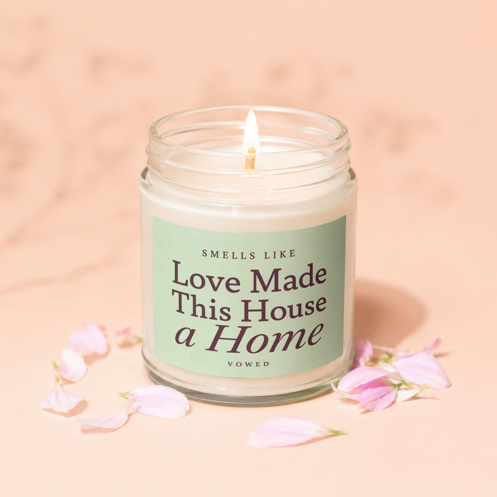 Glass candle with lit wick and light green label that reads "Smells Like Love Made This House a Home".