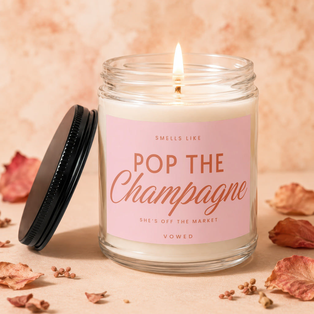 Glass jar candle with pink label reading "Smells Like Pop The Champagne She's Off The Market Vowed."