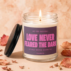 Lit candle in a glass jar with purple label reading "Love Never Feared the Dark".