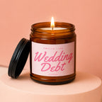 Lit candle in amber jar with "Smells Like Wedding Debt" label on pink background.