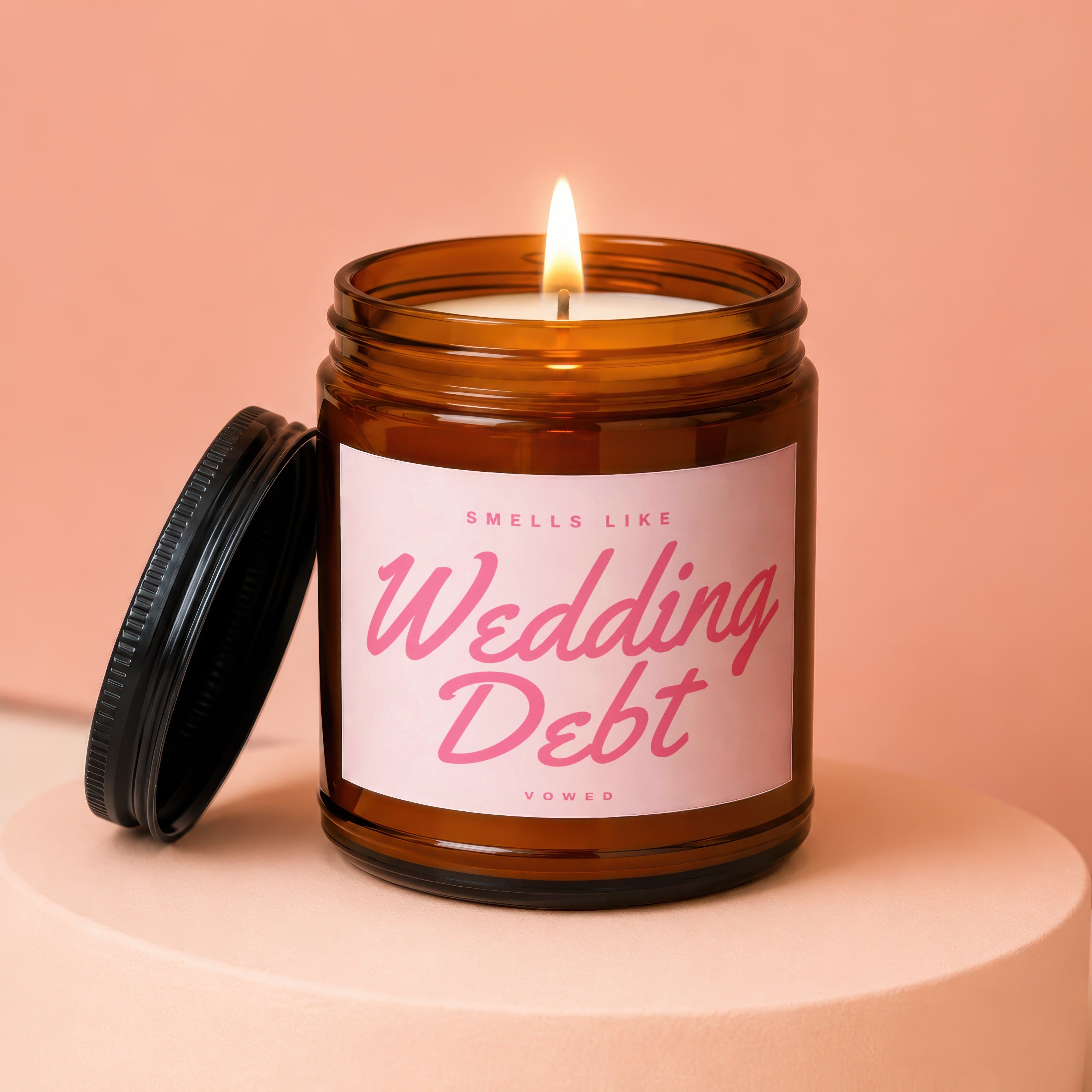 Lit candle in amber jar with "Smells Like Wedding Debt" label on pink background.