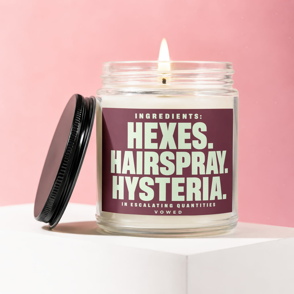 Glass candle with burning wick and lid, featuring label text: "Ingredients: Hexes. Hairspray. Hysteria. In Escalating Quantities. Vowed."